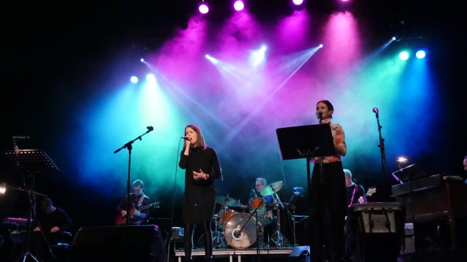 Personer står på mörk scen med strålkastare och sjunger. Ett band spelar musik i bakgrunden.