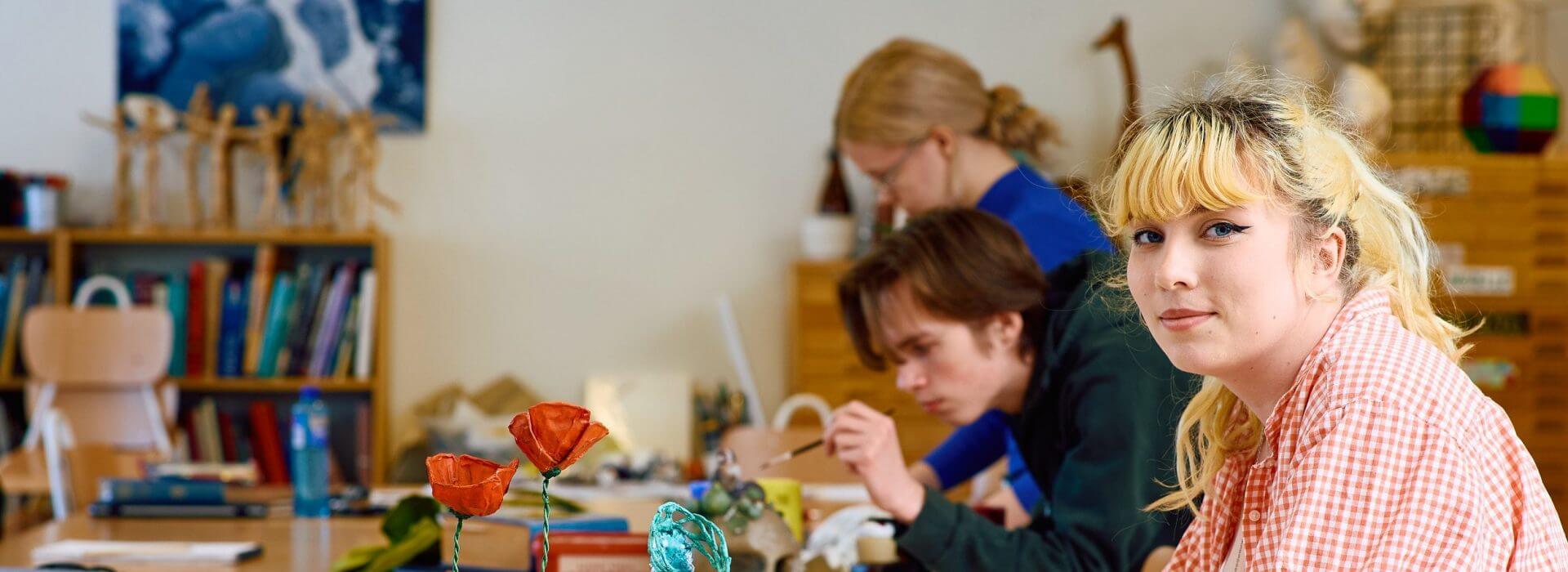 Kvinnlig elev sitter vid ett bord i en i bildsal med klasskamrater som jobbar på sina konstverk i bakgrunden